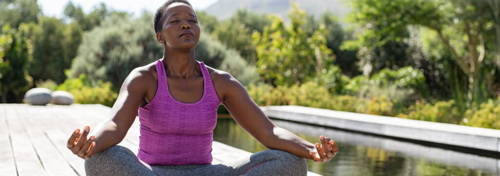 Mature African American woman sits outdoors in a lotus postion meditating next to a pond.